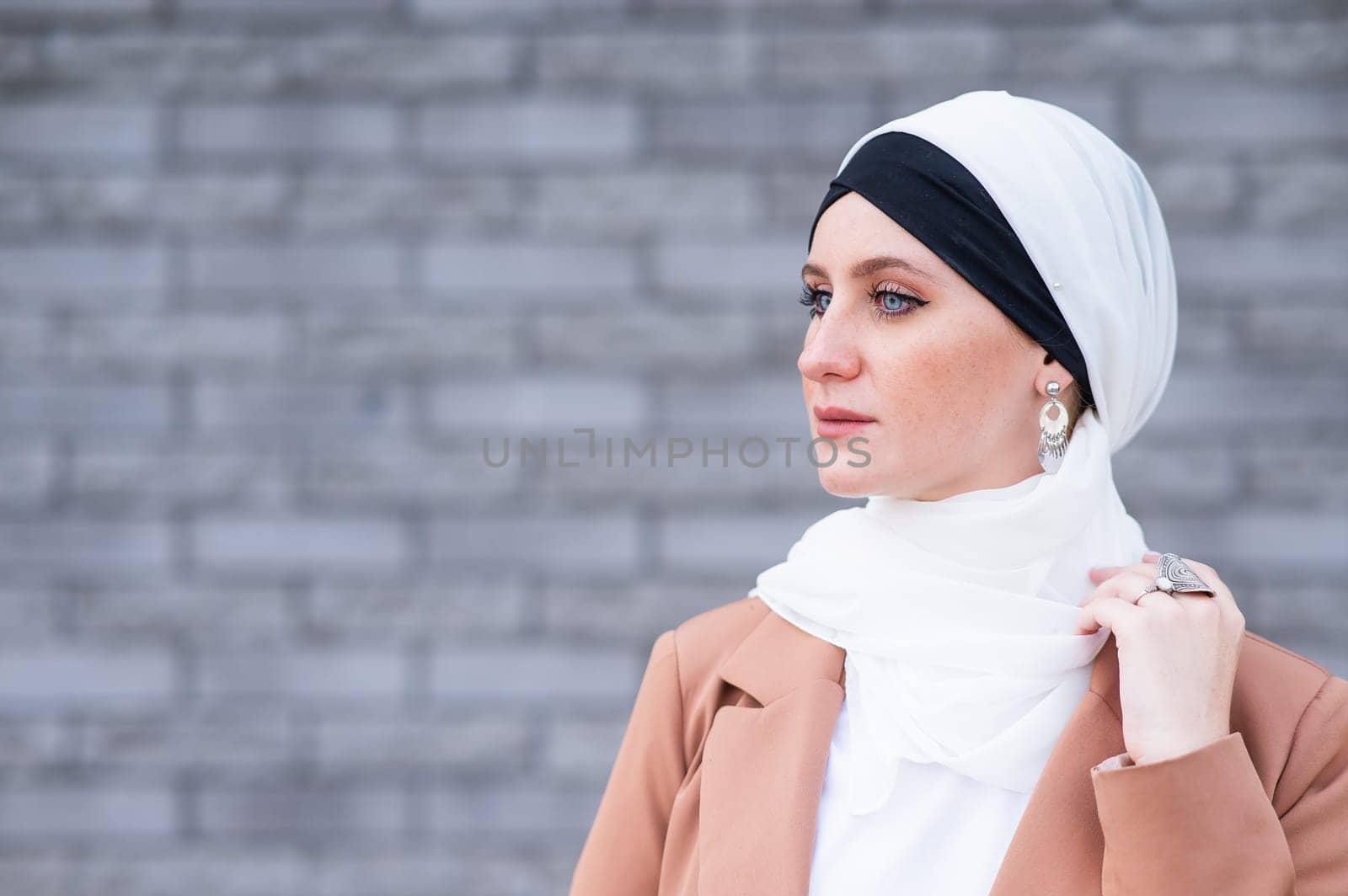 Portrait of a young blue-eyed woman in a hijab against a gray brick wall