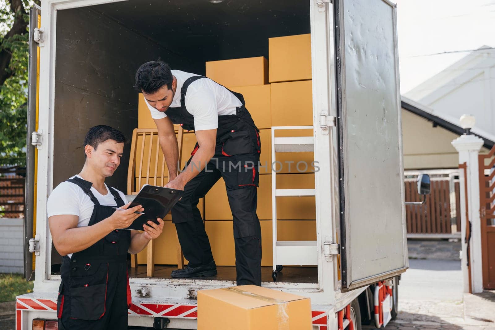 Uniformed workers unload cardboard boxes checking checklist with a clipboard at the truck. Reliable movers guarantee efficient moving and professional service. Moving day concept by Sorapop