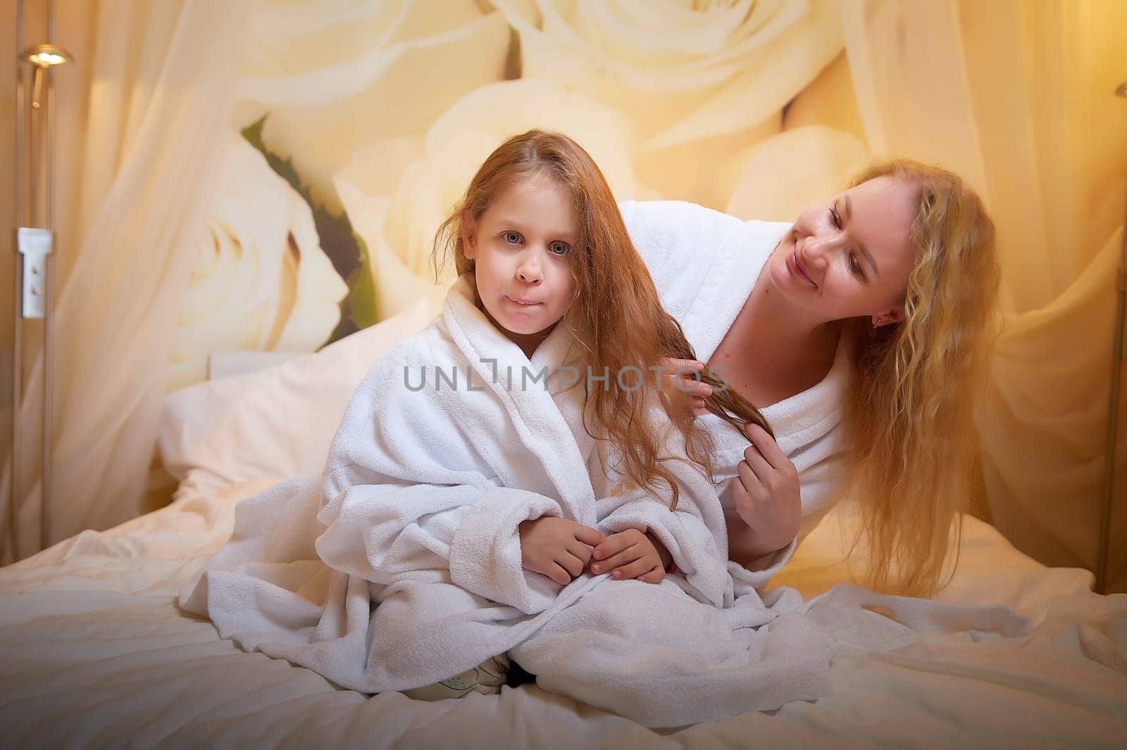 Mother and daughter happily relax and fun together on bed in bedroom. The concept of tenderness between mom and girl. Mom braids the girl's hair