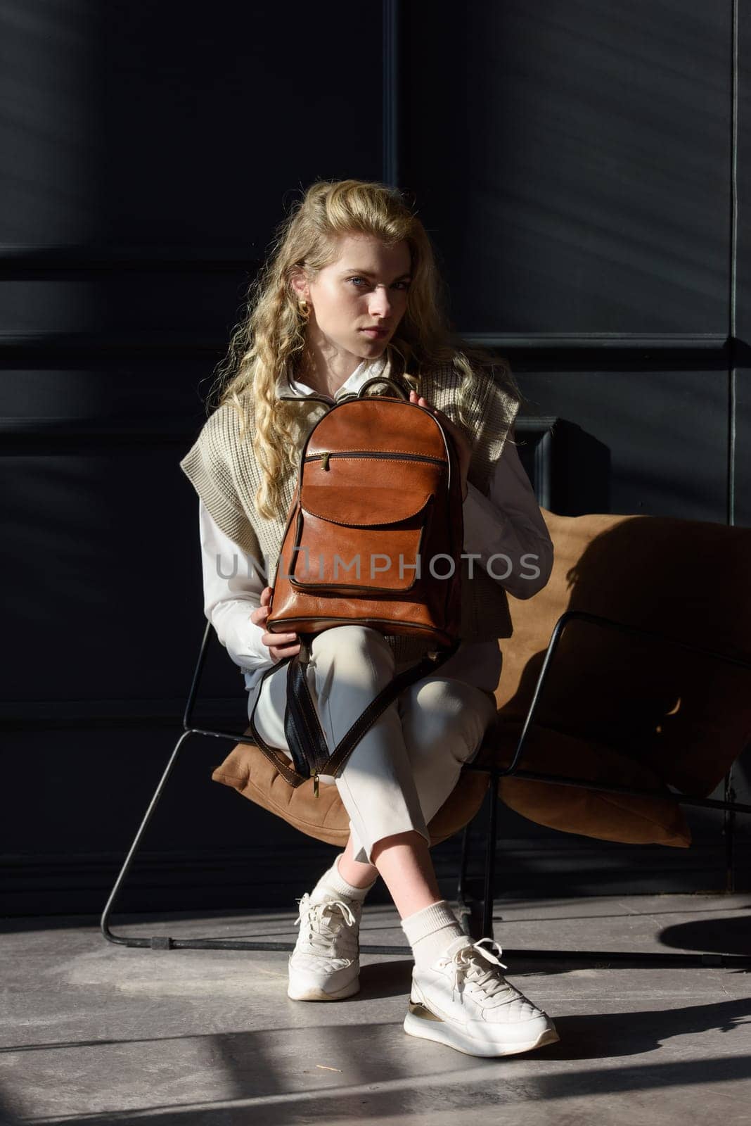 A woman holds leather backpack. Model wearing stylish knitted vest, white shirt and classic trousers.