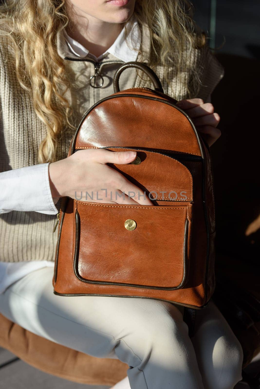 A woman holds leather backpack. Model wearing stylish knitted vest, white shirt and classic trousers.