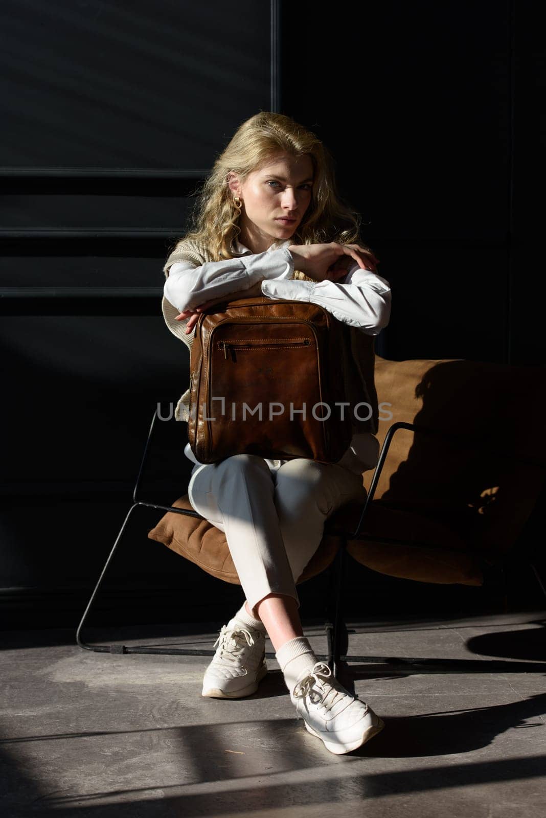 A woman holds leather backpack. Model wearing stylish knitted vest, white shirt and classic trousers.