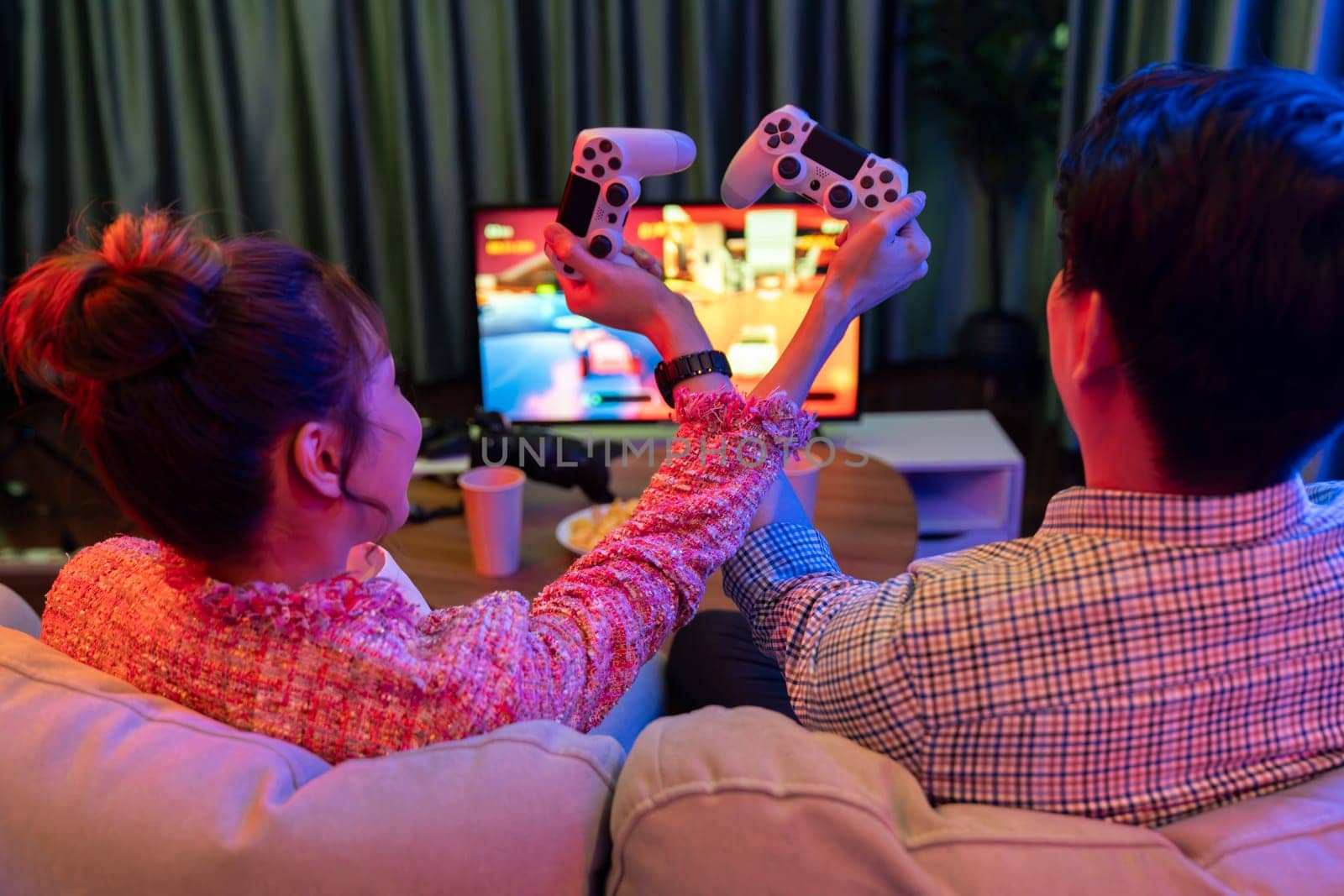 Couple gamer with hands cross of joystick playing fighting competition video game together on tv screen at back side view, sitting sofa at neon light color living room at modern comfy home. Infobahn.