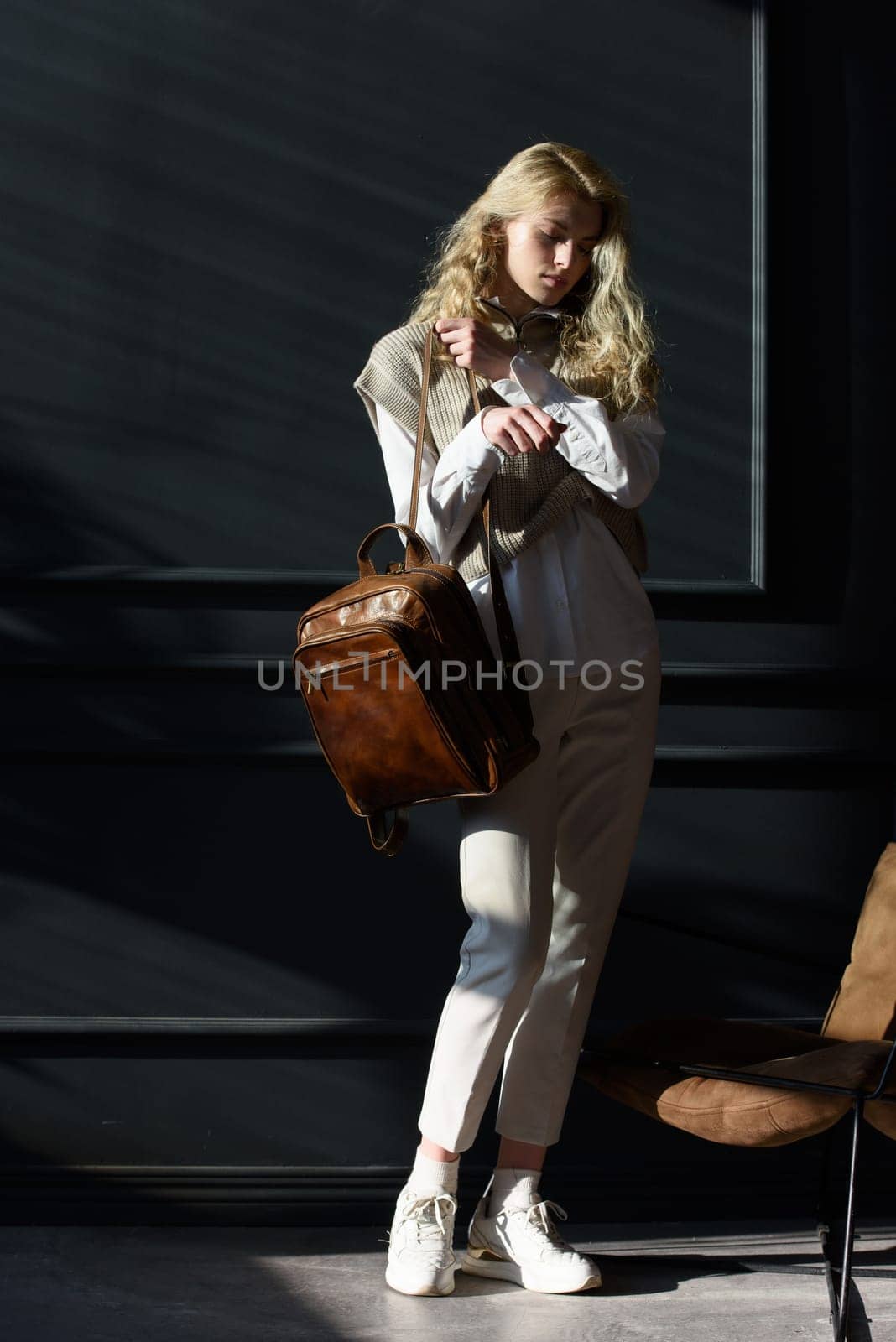 A woman holds brown leather backpack. Model wearing stylish knitted vest, white shirt and classic trousers.