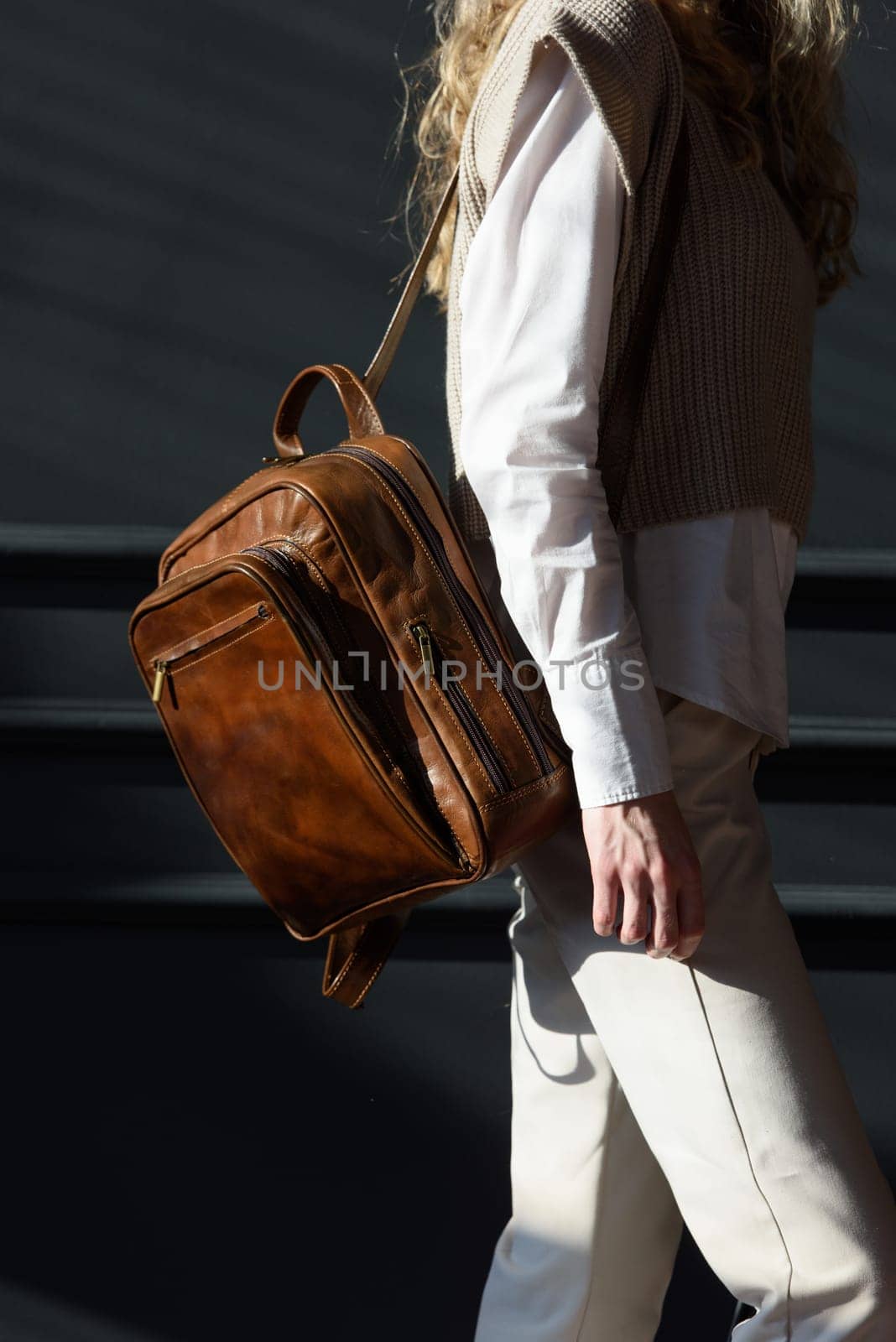 A woman holds brown leather backpack. Model wearing stylish knitted vest, white shirt and classic trousers.