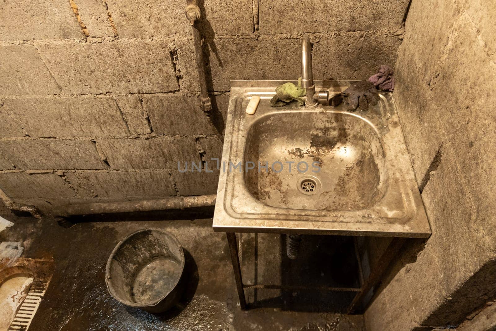 Dirty toilet room stainless steel sink with a bucket on the floor, uncoated concrete flooring and cinder brick walls. by z1b