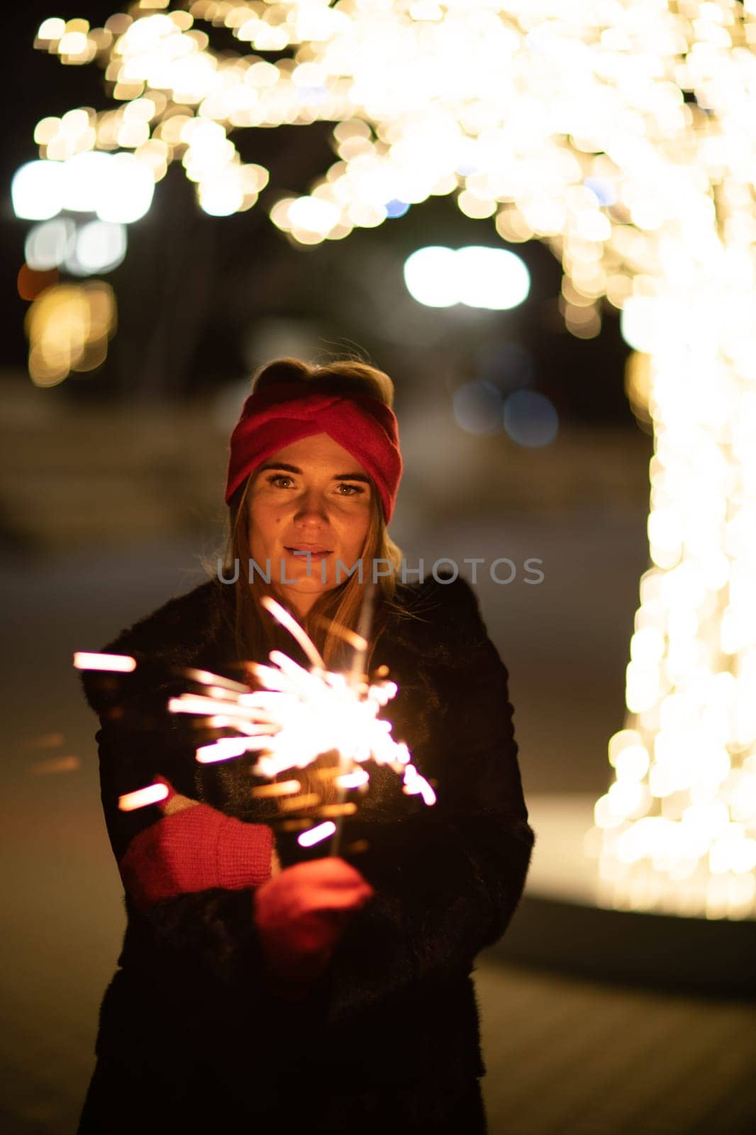 Woman holding sparkler night while celebrating Christmas outside. Dressed in a fur coat and a red headband. Blurred christmas decorations in the background. Selective focus by Matiunina