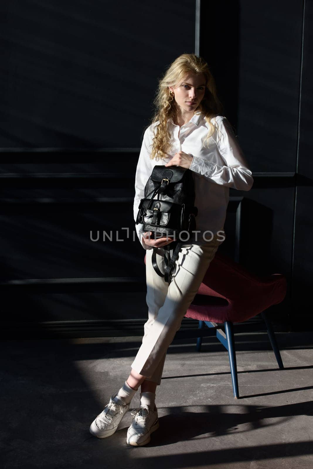 A woman holds black leather backpack. Model wearing stylish knitted vest, white shirt and classic trousers.