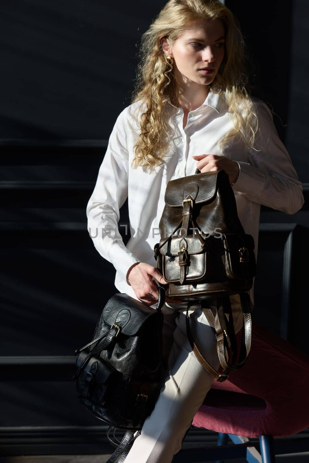 A woman holds two brown leather backpacks. Model wearing stylish knitted vest, white shirt and classic trousers.