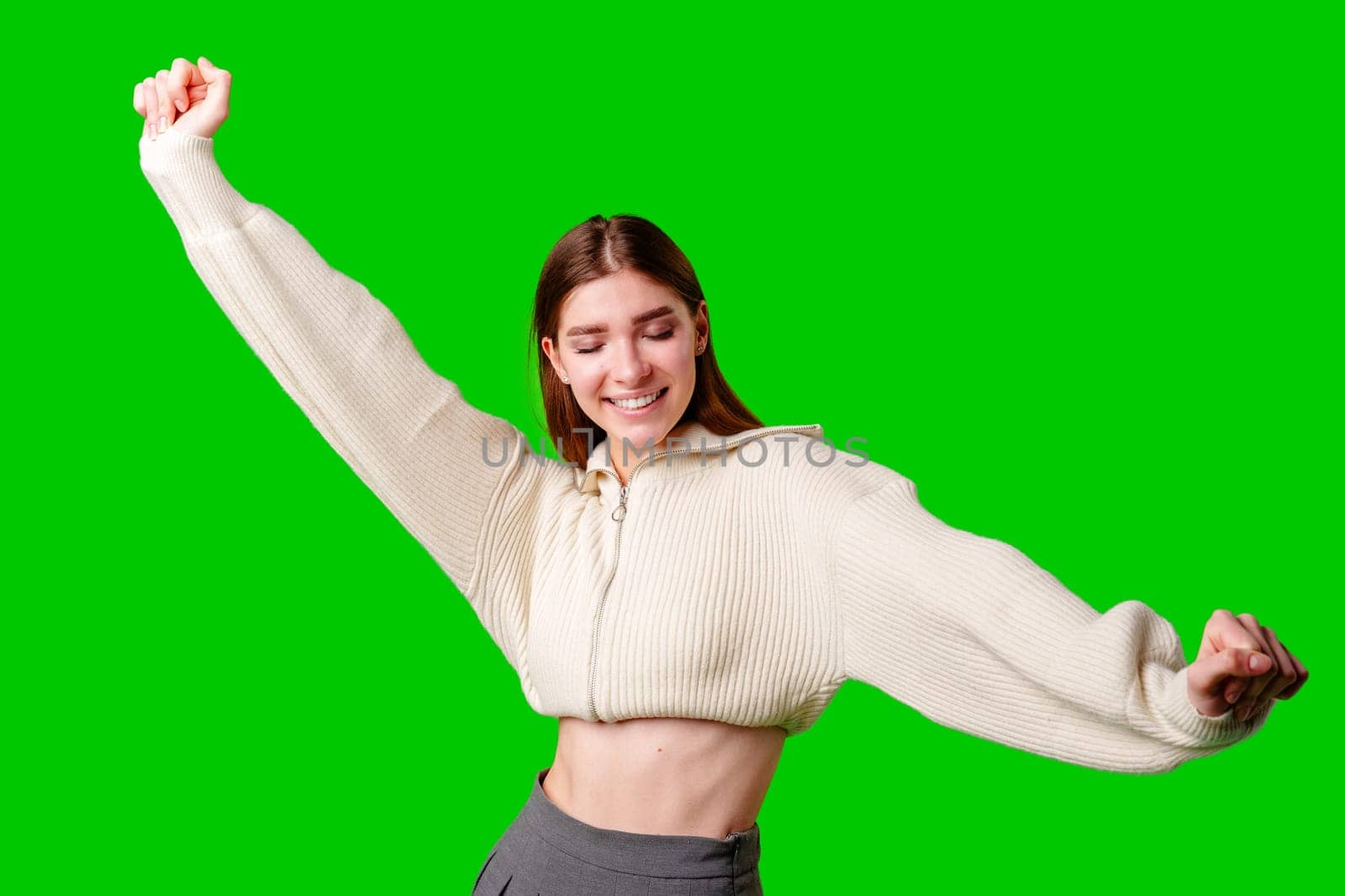 A young woman is captured in a moment of jubilation, her arms raised in victory and a beaming smile on her face, set against a vivid green background which indicates the use of a green screen for post-production editing.