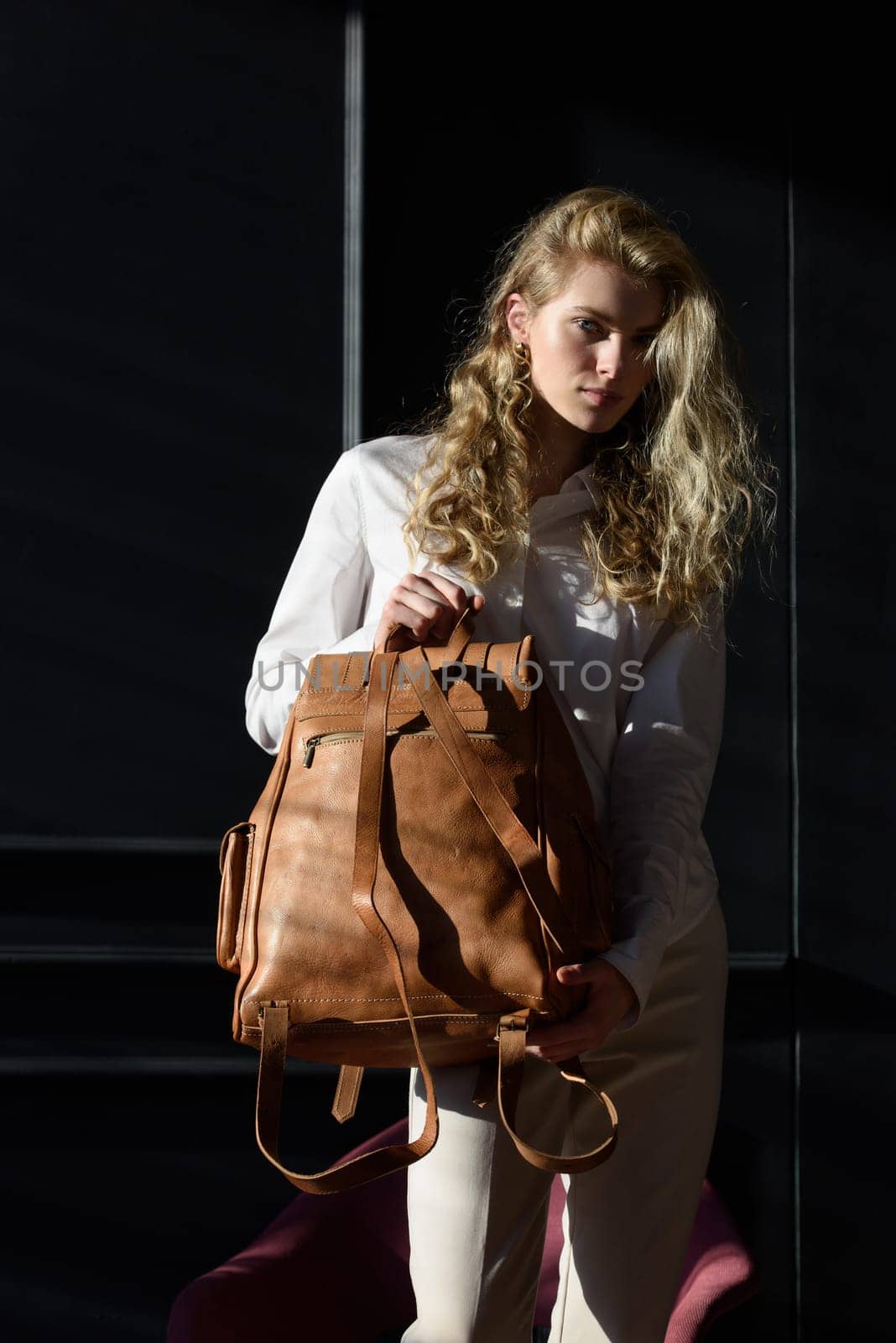 A woman holds brown leather backpack. Model wearing stylish knitted vest, white shirt and classic trousers.