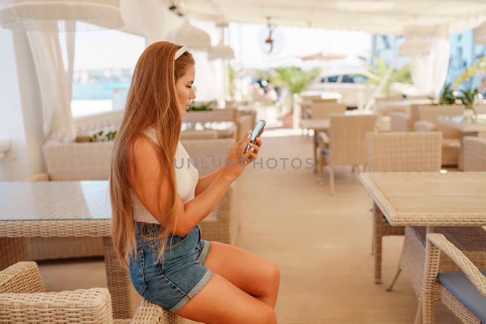 Cafe woman sitting at a table with a cell phone in her hand. She is looking at the screen and she is focused on her phone. The scene takes place in a restaurant or cafe, with multiple chairs