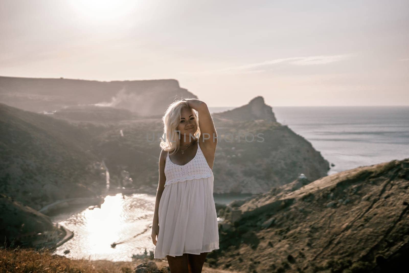 A blonde woman stands on a hill overlooking the ocean. She is wearing a white dress and she is enjoying the view. by Matiunina