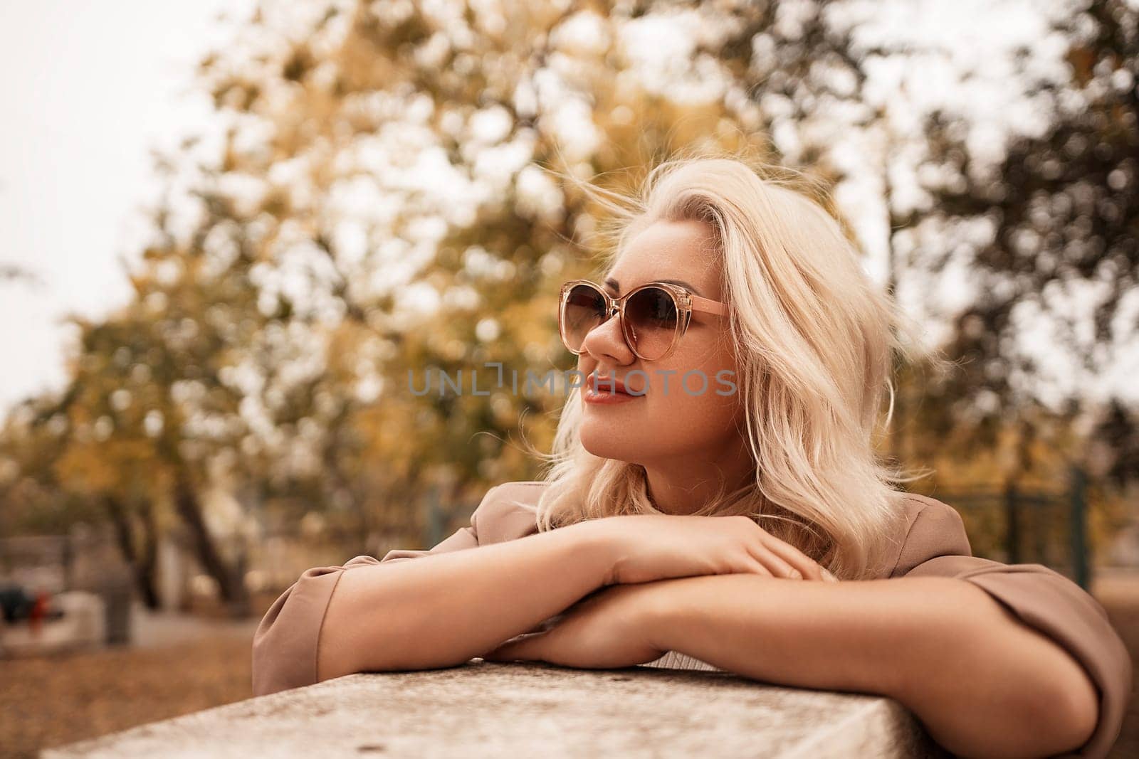 A blonde woman wearing sunglasses is sitting on a bench in a park. She is looking at the camera with a smile on her face