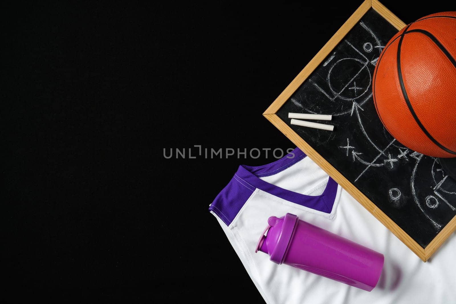 A basketball, along with a strategic play diagrammed on a chalkboard, rests near a folded jersey and a sports bottle. The equipment is arranged on a dark surface, suggesting preparation for a game or a coaching session in progress.