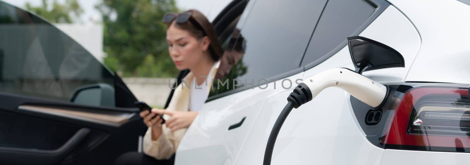 Young woman use smartphone to pay for electricity at public EV car charging station green city park. Modern environmental and sustainable urban lifestyle with EV vehicle. Panorama Expedient