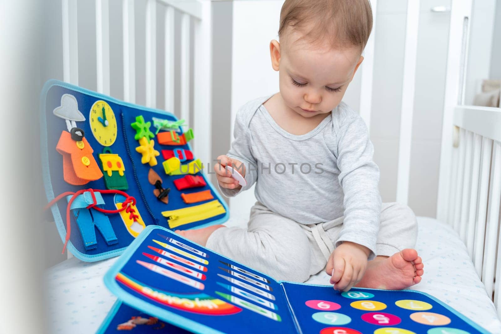 Little baby girl playing with busy book while sitting in crib. Concept of smart quiet books and modern educational toys