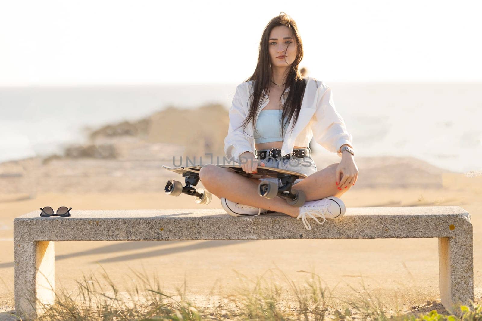 A woman is sitting on a bench with a skateboard in her hand. She is wearing a white shirt and blue shorts. The scene is set on a beach, with the ocean in the background