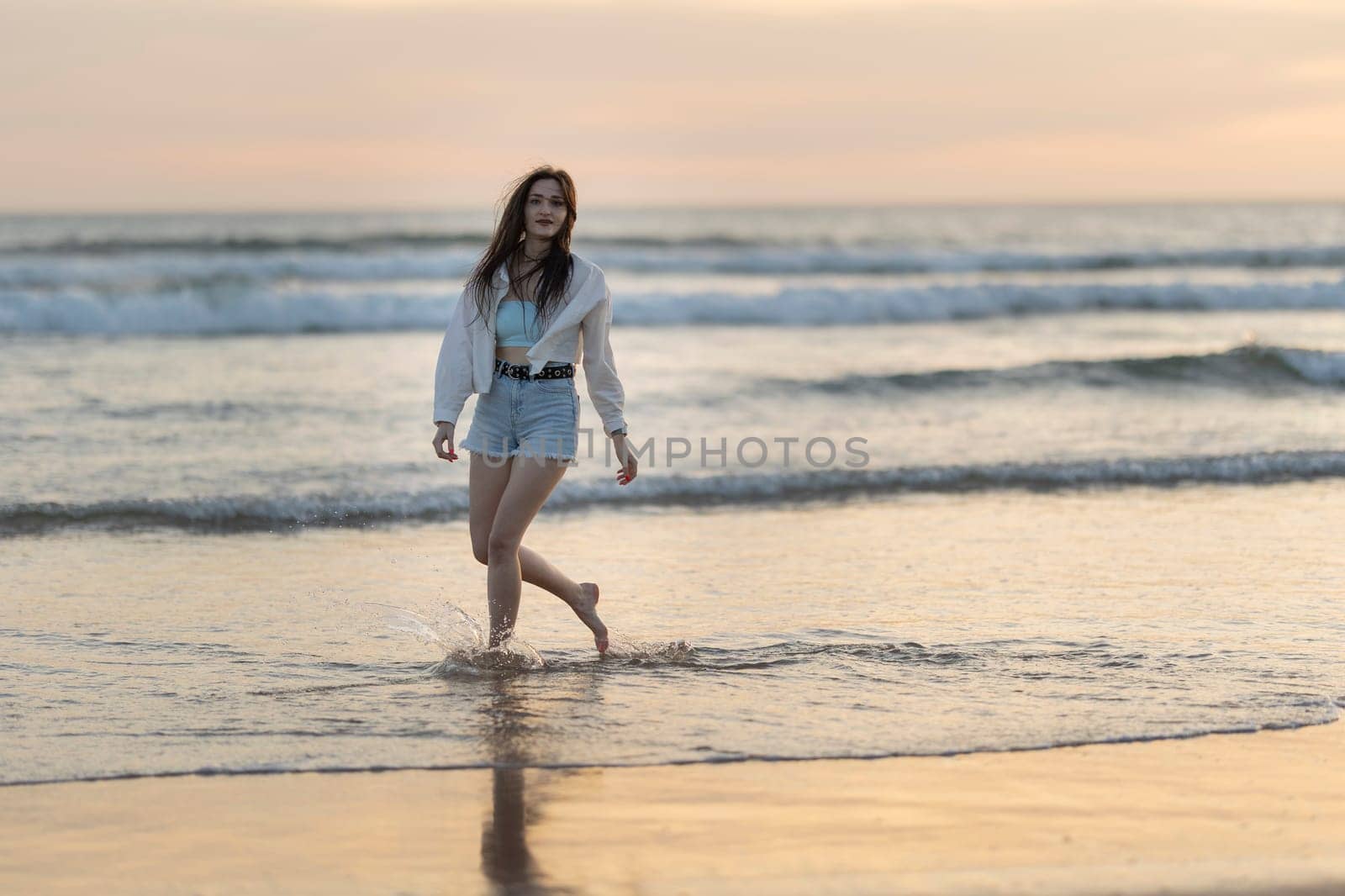 A woman is walking on the beach in the water. She is wearing a white shirt and blue shorts. The water is calm and the sky is orange