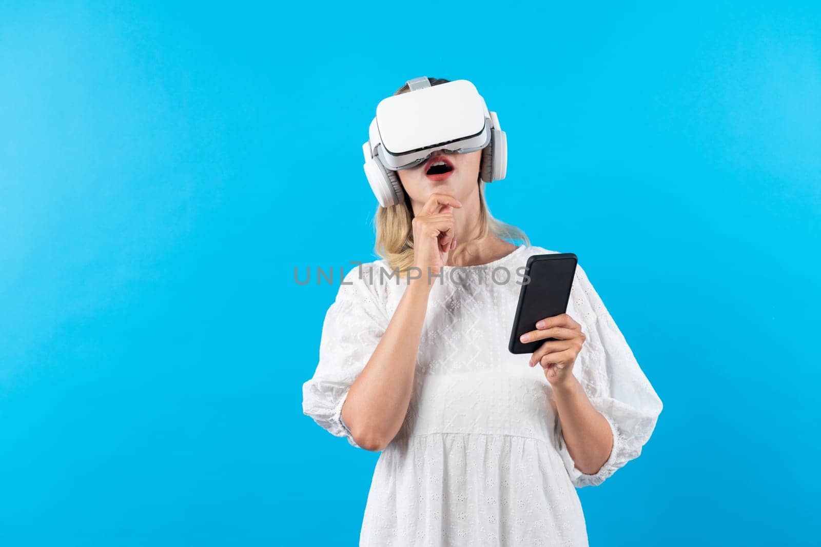 Caucasian girl wearing VR glasses while holding phone and pointing at view. Happy woman surprised while looking around to explore visual reality world or metaverse. Technology innovation. Contraption.