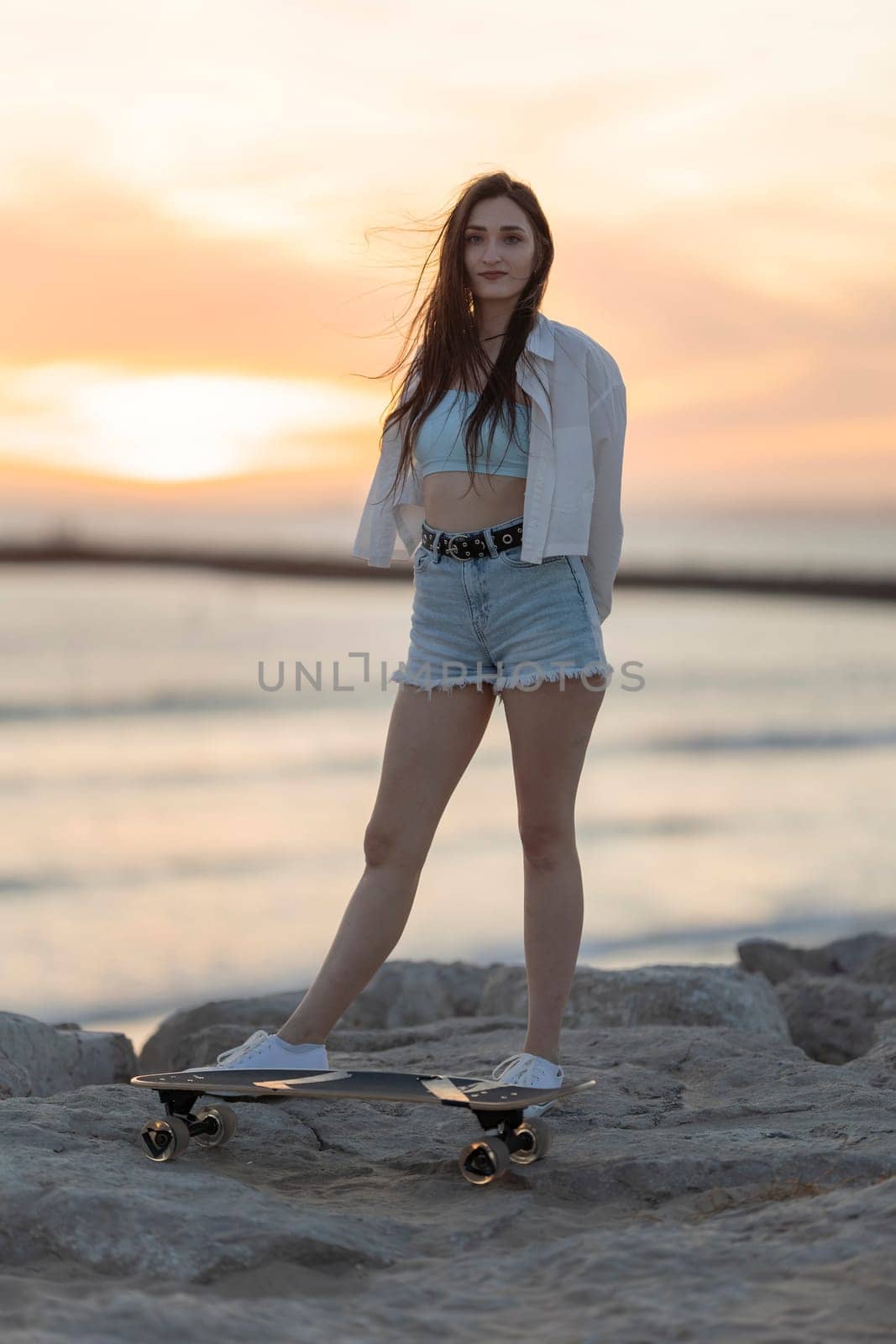 A woman is standing on a skateboard on a beach. The sky is orange and the water is calm