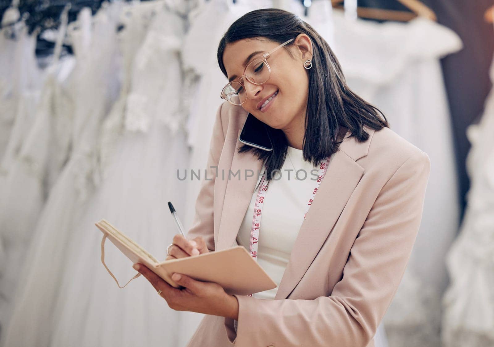 Notebook, phone call and wedding dress with woman in studio for communication or planning. Style, writing and fashion designer with tape measure talking on mobile in workshop for fitting appointment.