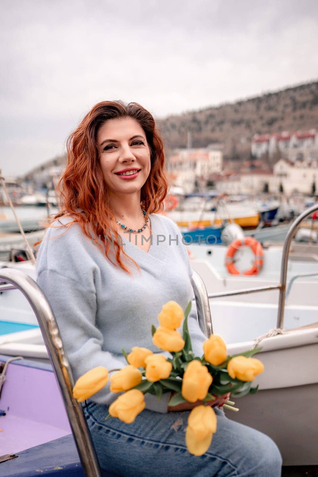 Woman holds yellow tulips in harbor with boats docked in the background., overcast day, yellow sweater, mountains.