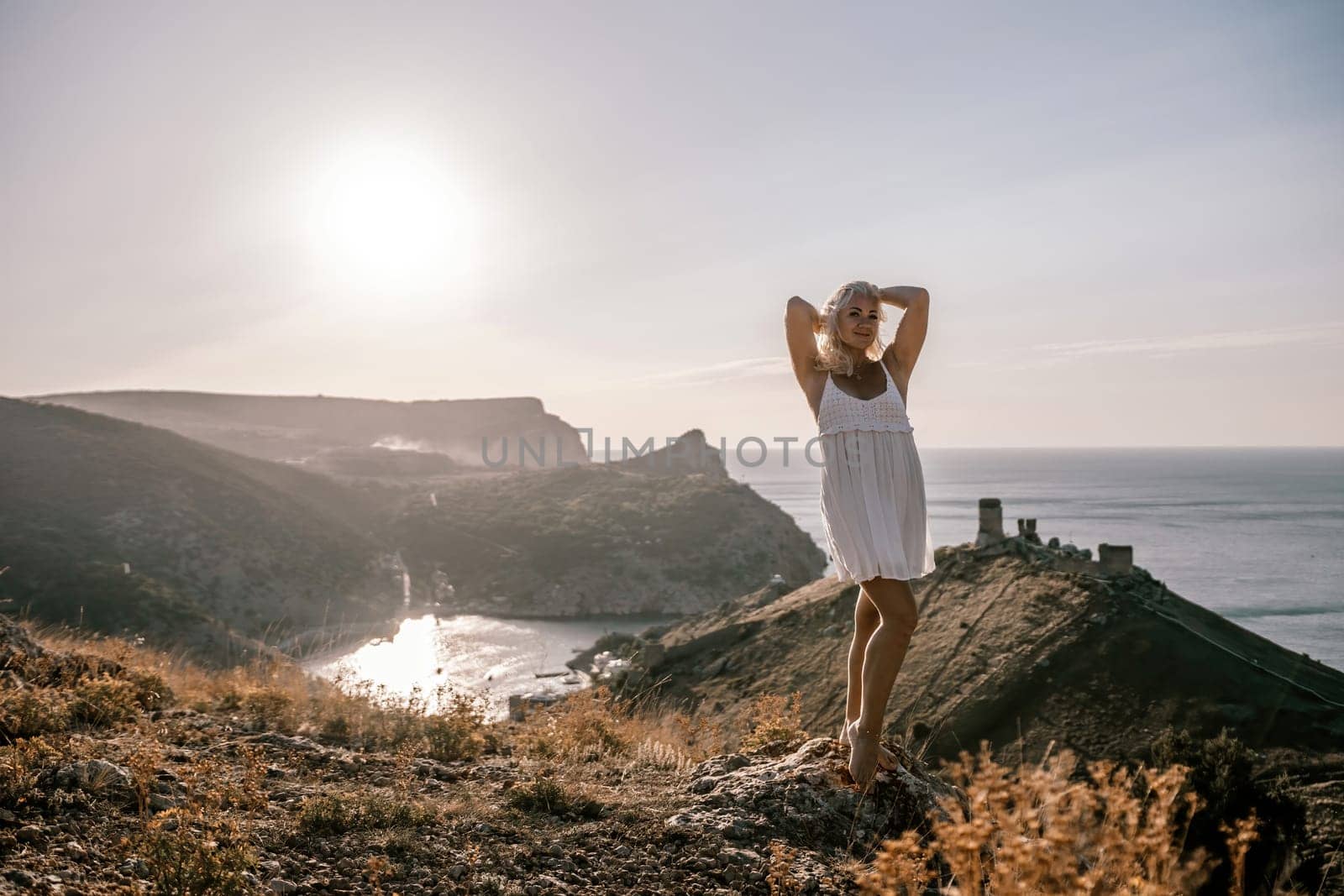 A blonde woman stands on a hill overlooking the ocean. She is wearing a white dress and she is enjoying the view. by Matiunina