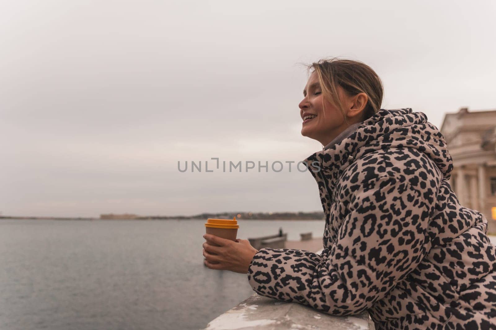 A woman in a leopard print coat is holding a coffee cup and looking out at the water