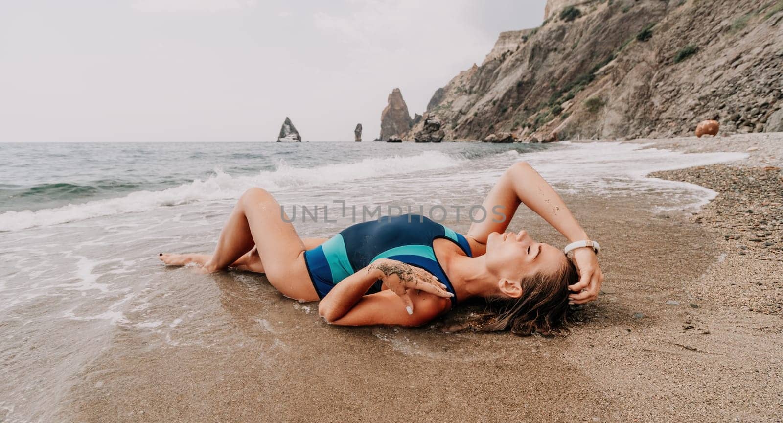 Woman summer travel sea. Happy tourist in blue bikini enjoy taking picture outdoors for memories. Woman traveler posing on the beach surrounded by volcanic mountains, sharing travel adventure journey by panophotograph