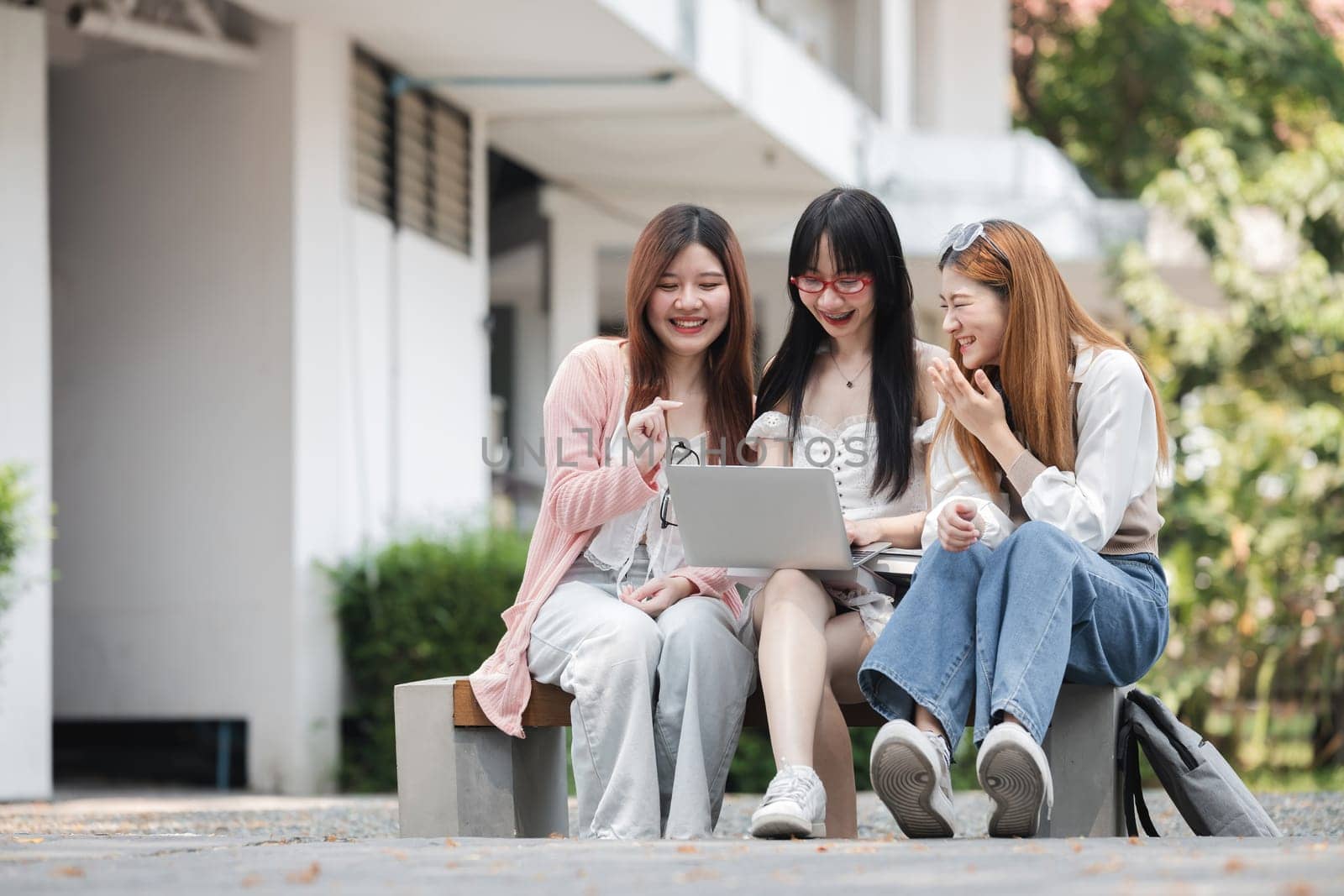 Group of happy young Asian college students sitting on a bench, looking at a laptop screen, discussing and brainstorming on their school project together..