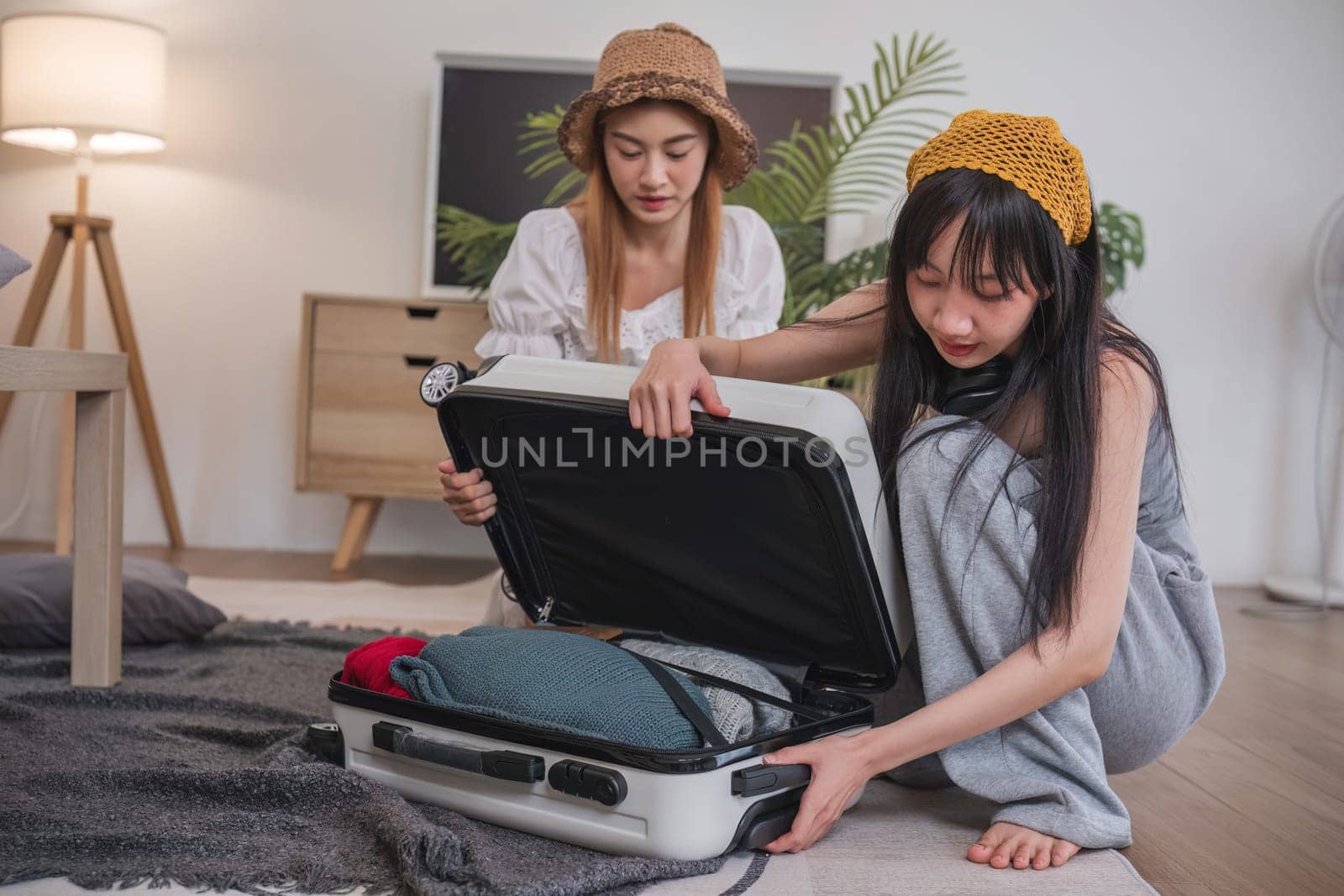 Two women are opening a suitcase in a living room. One of them is wearing a yellow hat