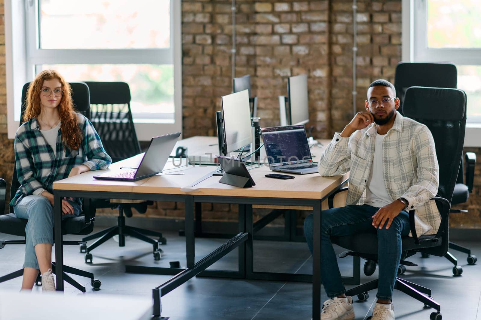 Group of colleagues, a woman with vibrant orange hair and a young African American businessman, sitting in a modern office space, symbolizing diverse collaboration and a dynamic work environment