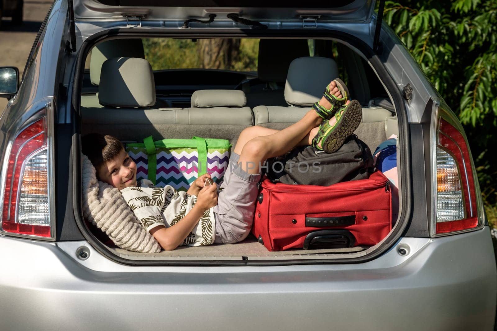 Cute little boy laying on the back of the bags and baggage in the car trunk ready to go on vacation with happy expression. Kid resting playing on smartphone by Kobysh