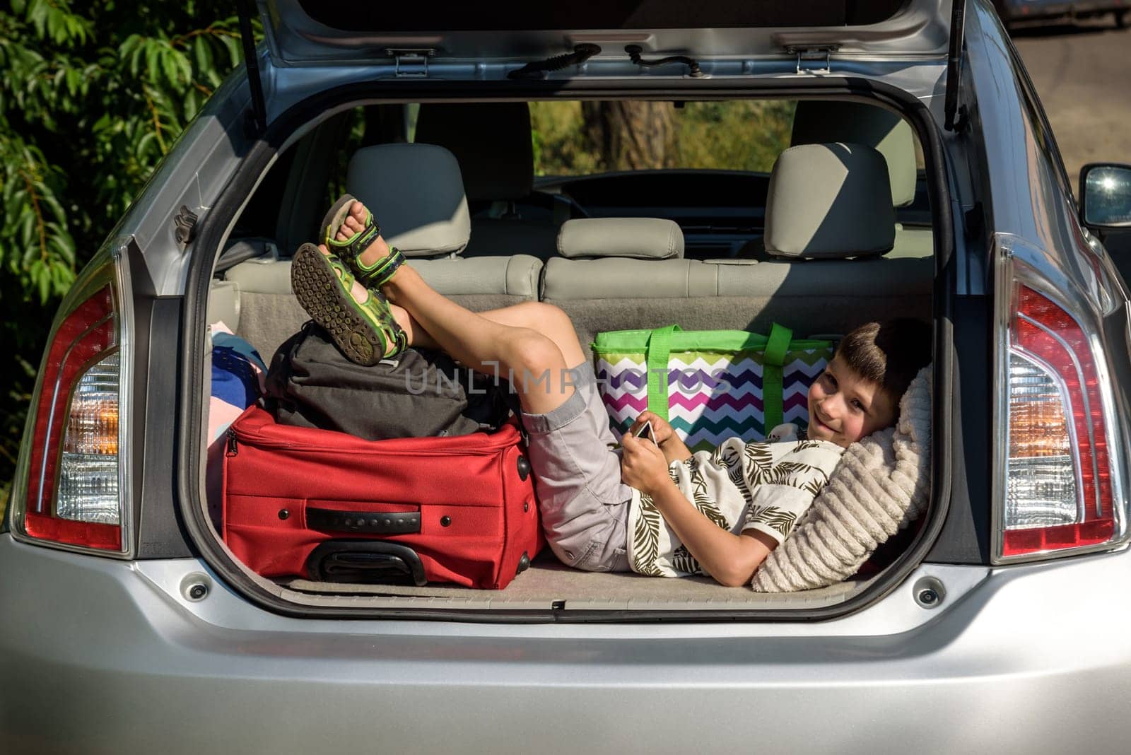 Cute little boy laying on the back of the bags and baggage in the car trunk ready to go on vacation with happy expression. Kid resting playing on smartphone by Kobysh