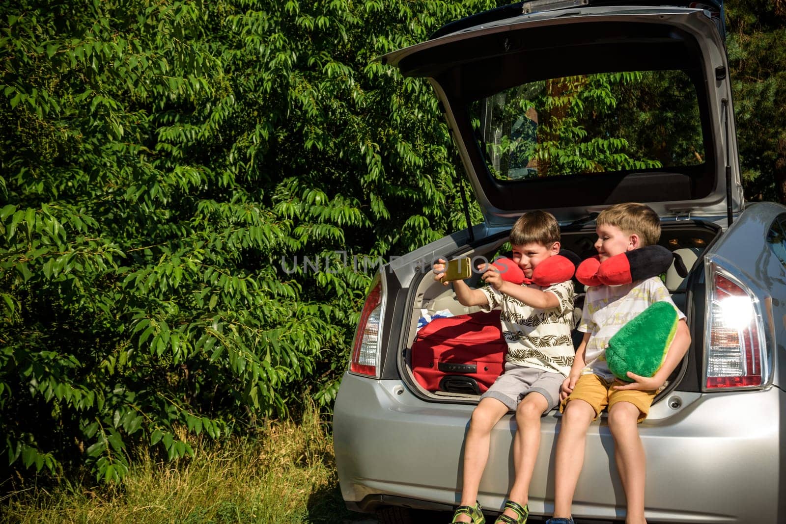 Two adorable little kids boy sitting in car trunk just before leaving for summer vacation. Sibling brothers making selfie on smartphone. Happy family going on long journey.