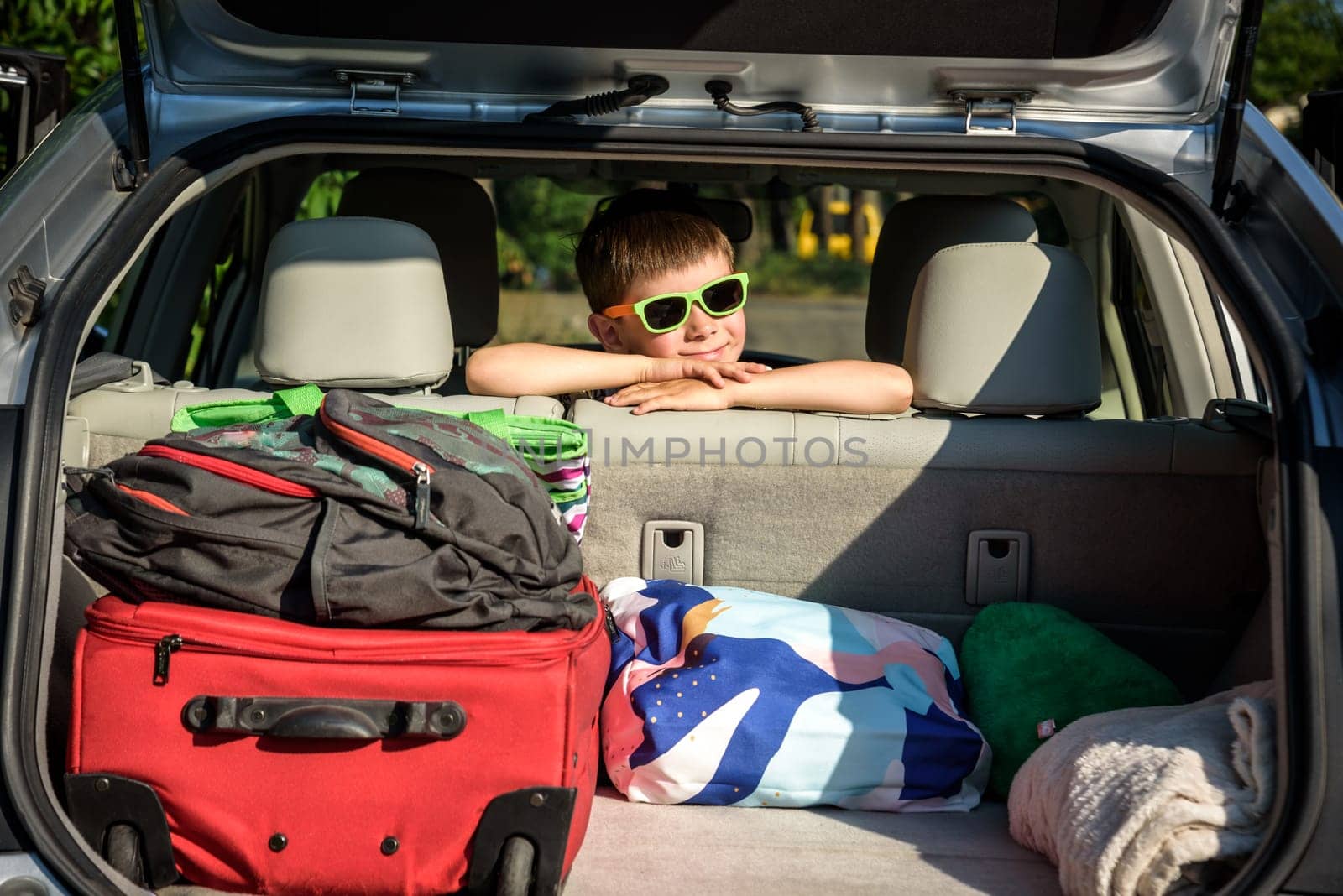 Adorable kid boy wearing sunglasses sitting in car trunk. Portrait of Happy child with open car boot while waiting for parent get ready for vocation. Family trip traveling by car concept by Kobysh