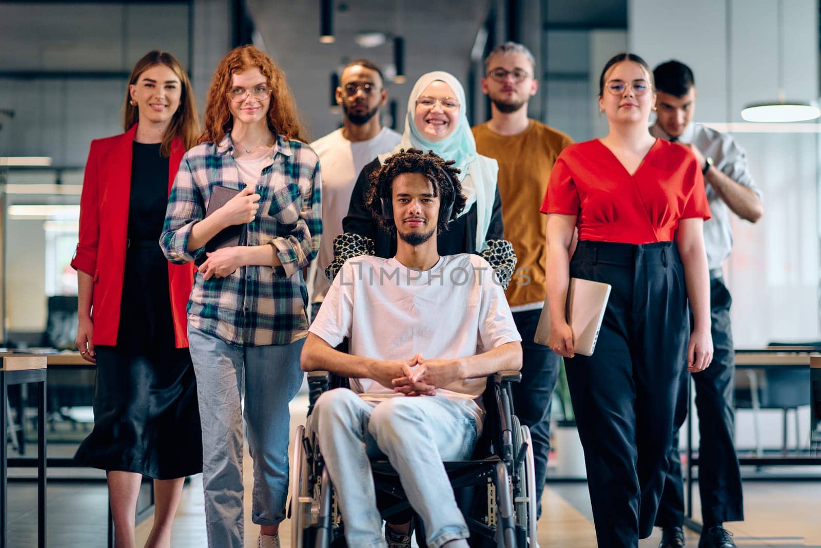 A diverse group of young business people walking a corridor in the glass-enclosed office of a modern startup, including a person in a wheelchair and a woman wearing a hijab.