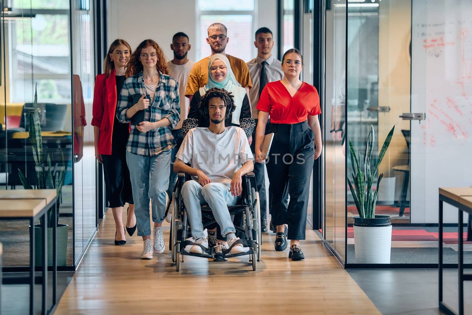 A diverse group of young business people congregates within a modern startup's glass-enclosed office, featuring inclusivity with a person in a wheelchair, an African American young man, and a hijab muslim woman