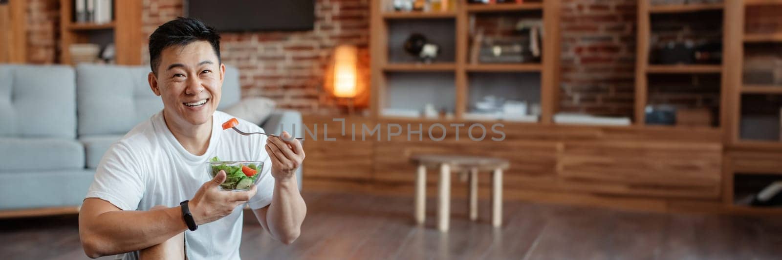 Healthy lifestyle concept. Happy middle aged asian man eating fresh vegetable salad after home workout and smiling at camera. Mature male keeping weight loss diet, panorama with copy space