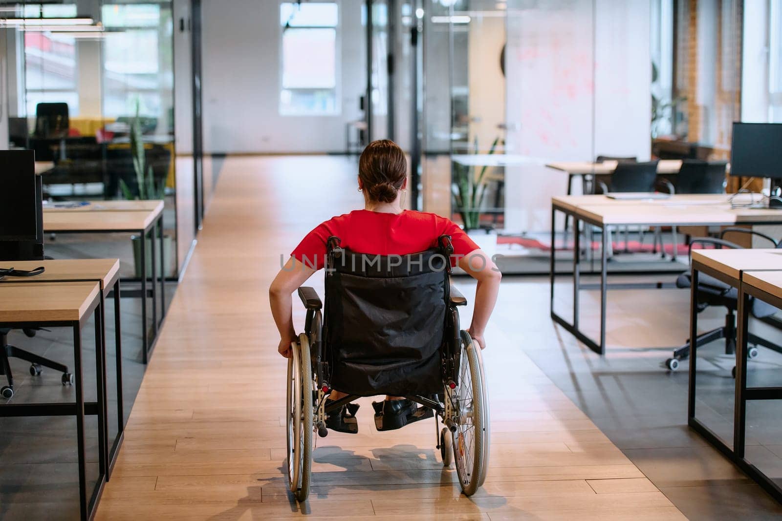 A modern young businesswoman in a wheelchair is surrounded by an inclusive workspace with glass-walled offices, embodying determination and innovation in the business world.