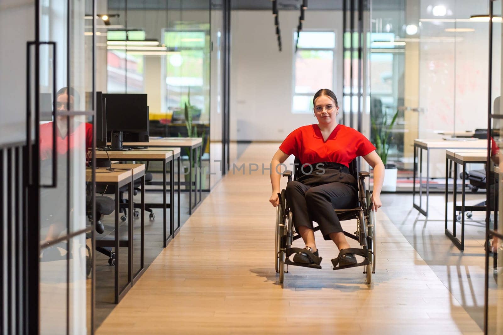 A modern young businesswoman in a wheelchair is surrounded by an inclusive workspace with glass-walled offices, embodying determination and innovation in the business world.