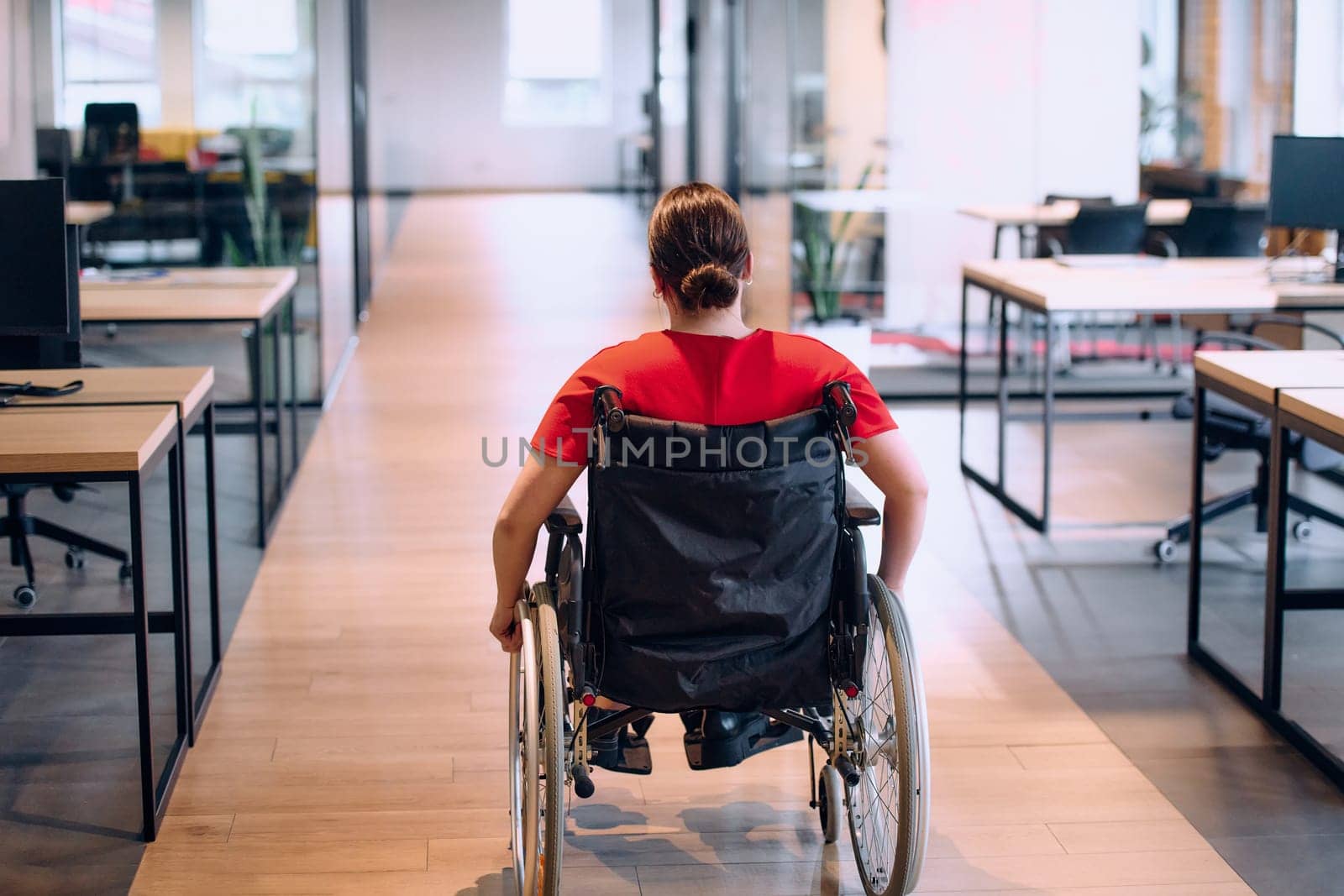 A modern young businesswoman in a wheelchair is surrounded by an inclusive workspace with glass-walled offices, embodying determination and innovation in the business world.