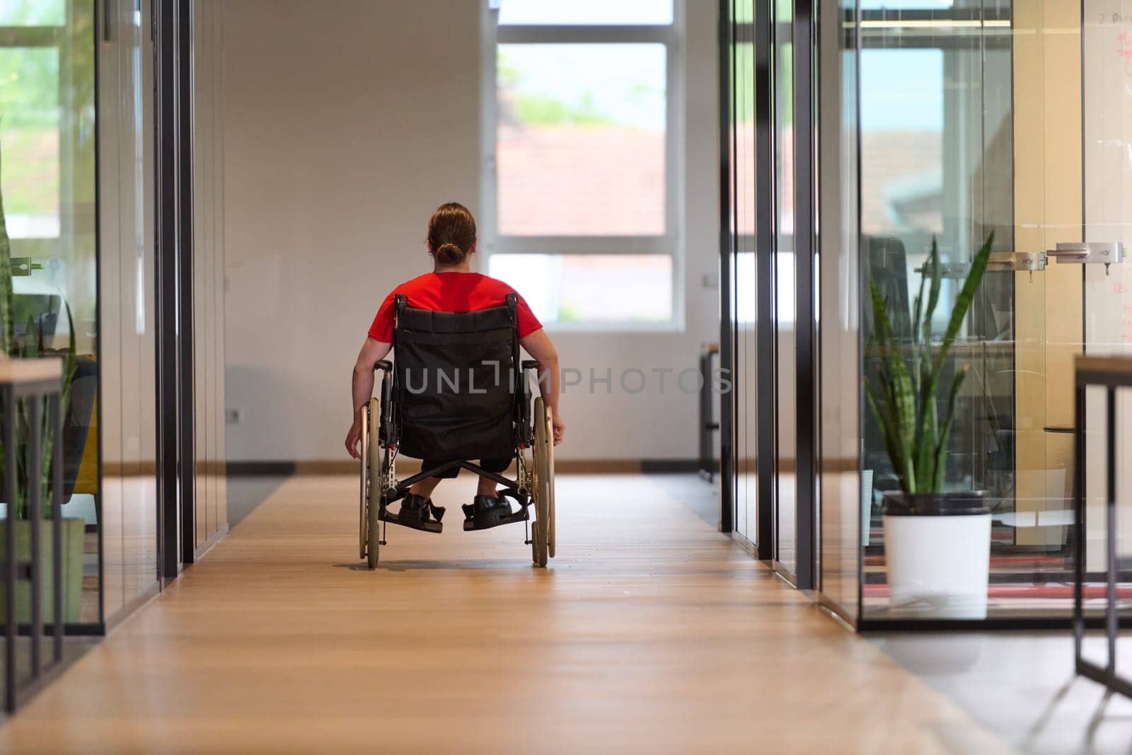 A modern young businesswoman in a wheelchair is surrounded by an inclusive workspace with glass-walled offices, embodying determination and innovation in the business world.