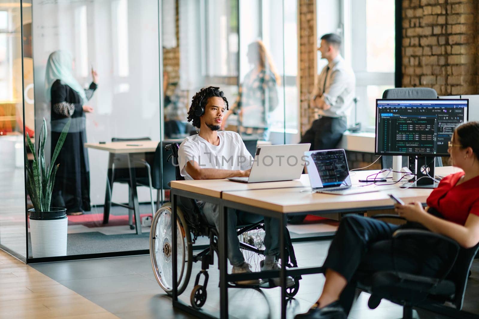 A young business group, including an African American businessman in a wheelchair, collaborates within a modern glass office, actively engaged around a computer and laptop, collectively solving diverse business challenges with determination by dotshock