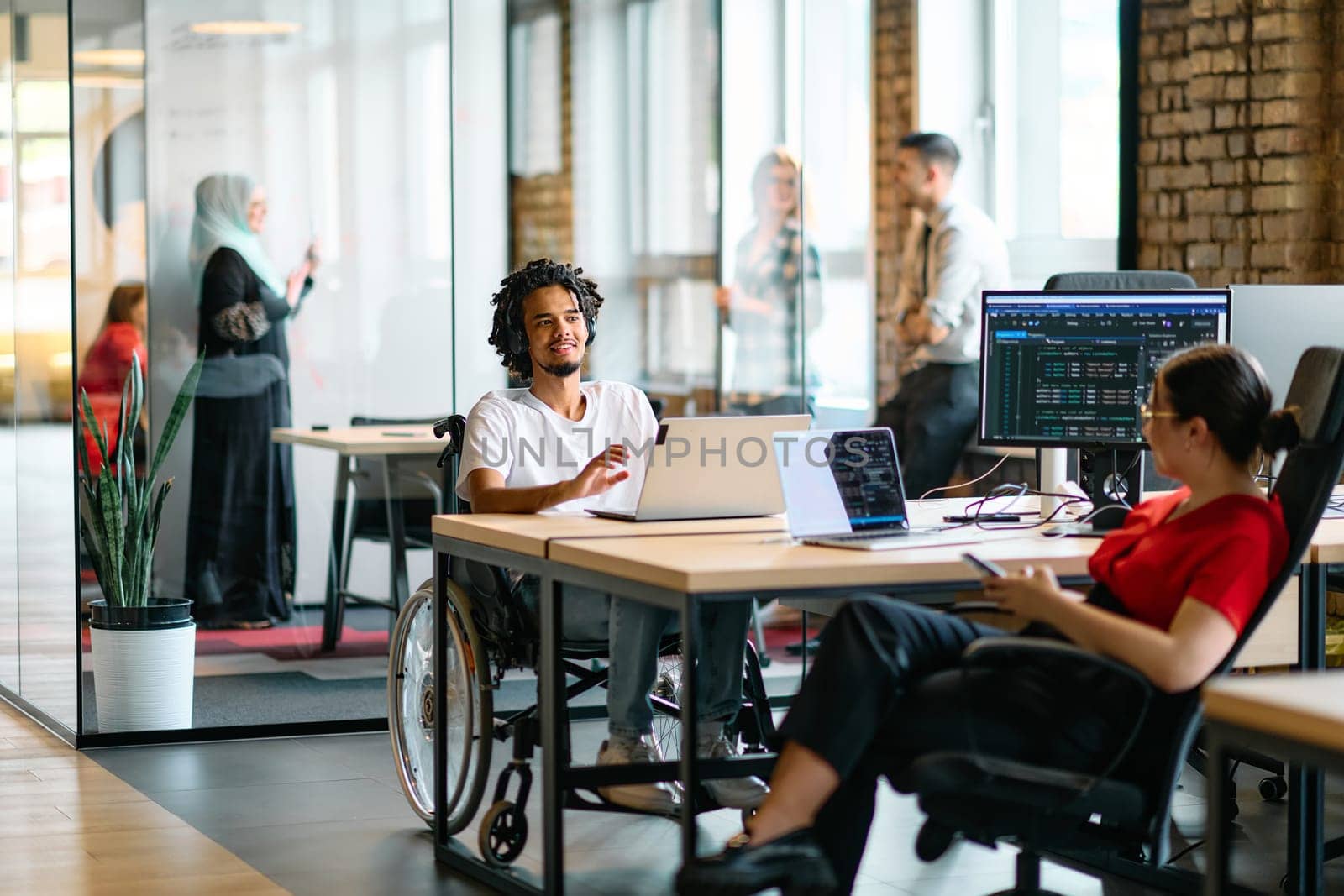 A young business group, including an African American businessman in a wheelchair, collaborates within a modern glass office, actively engaged around a computer and laptop, collectively solving diverse business challenges with determination.