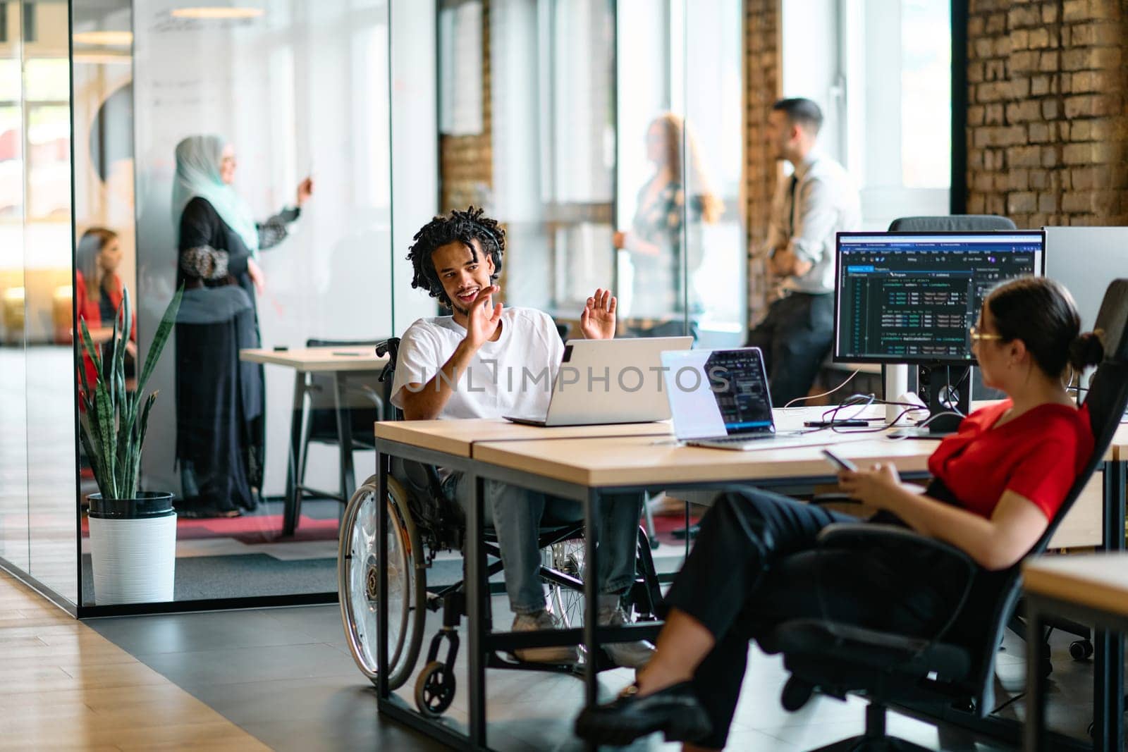 A young business group, including an African American businessman in a wheelchair, collaborates within a modern glass office, actively engaged around a computer and laptop, collectively solving diverse business challenges with determination by dotshock