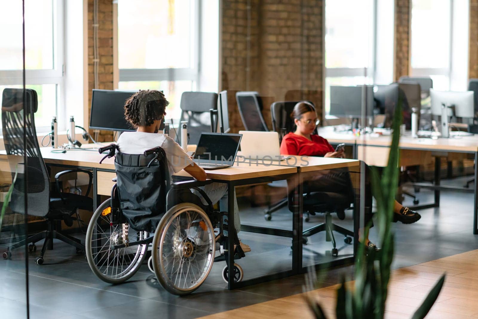 A young business group, including an African American businessman in a wheelchair, collaborates within a modern glass office, actively engaged around a computer and laptop, collectively solving diverse business challenges with determination.