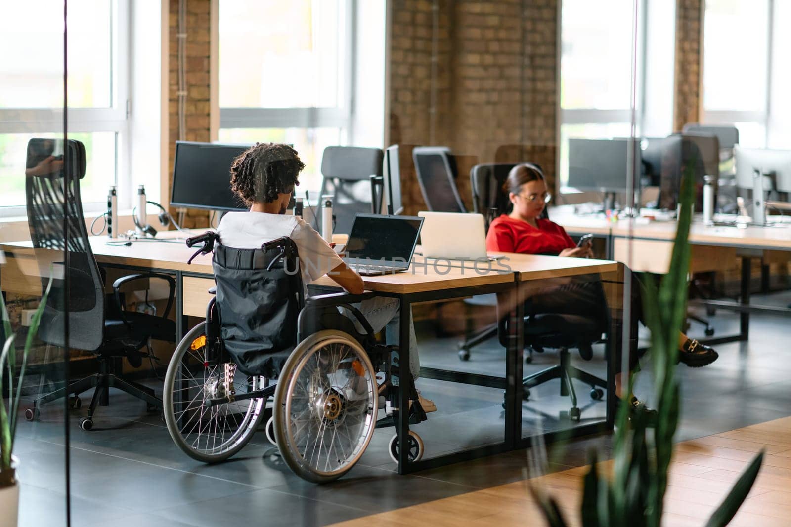 A young business group, including an African American businessman in a wheelchair, collaborates within a modern glass office, actively engaged around a computer and laptop, collectively solving diverse business challenges with determination.