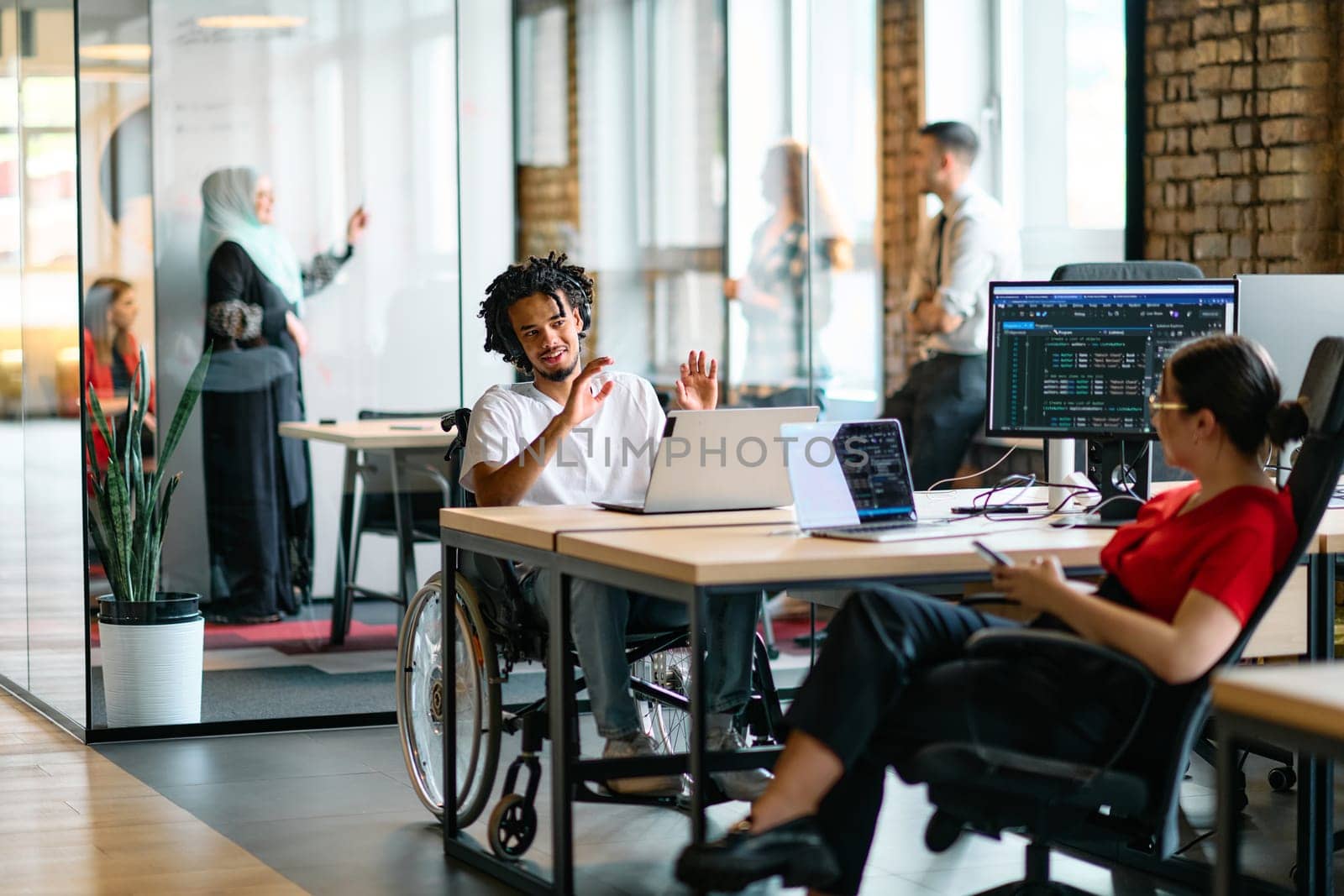 A young business group, including an African American businessman in a wheelchair, collaborates within a modern glass office, actively engaged around a computer and laptop, collectively solving diverse business challenges with determination by dotshock